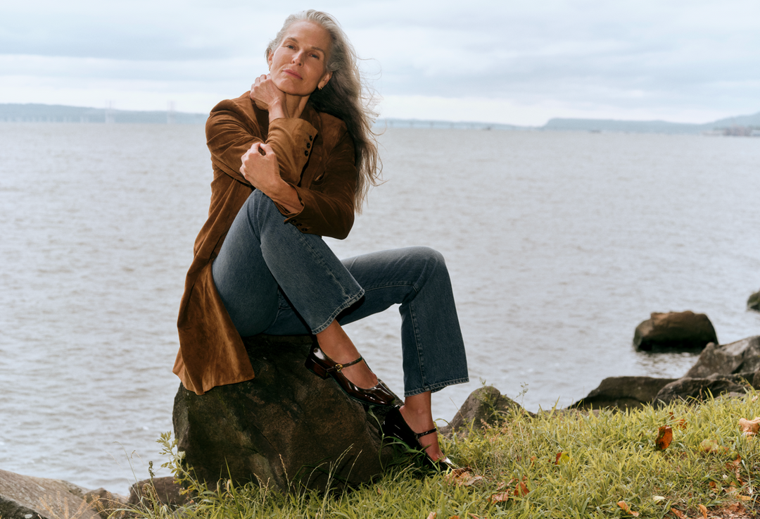 A woman sits on a large rock by the water, wearing a brown suede-like coat, blue jeans, and black strappy heels. The background shows a calm body of water under an overcast sky.
