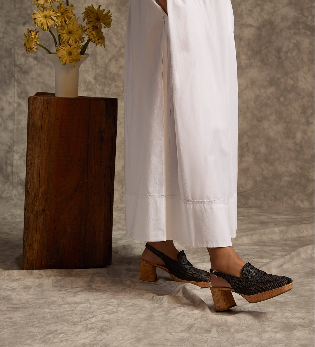Person wearing black shoes with wooden soles standing next to a wooden vase with yellow flowers on a textured floor.